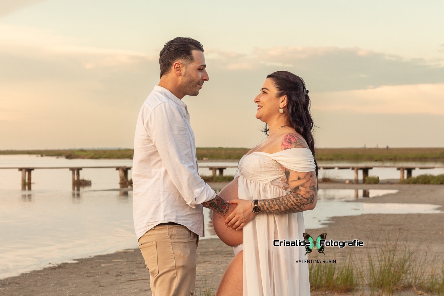 Mamma incinta di profilo in abito bianco guarda il marito sorridente, mani sulla pancia, sfondo spiaggia con mare, ciuffi d'erba e pontile