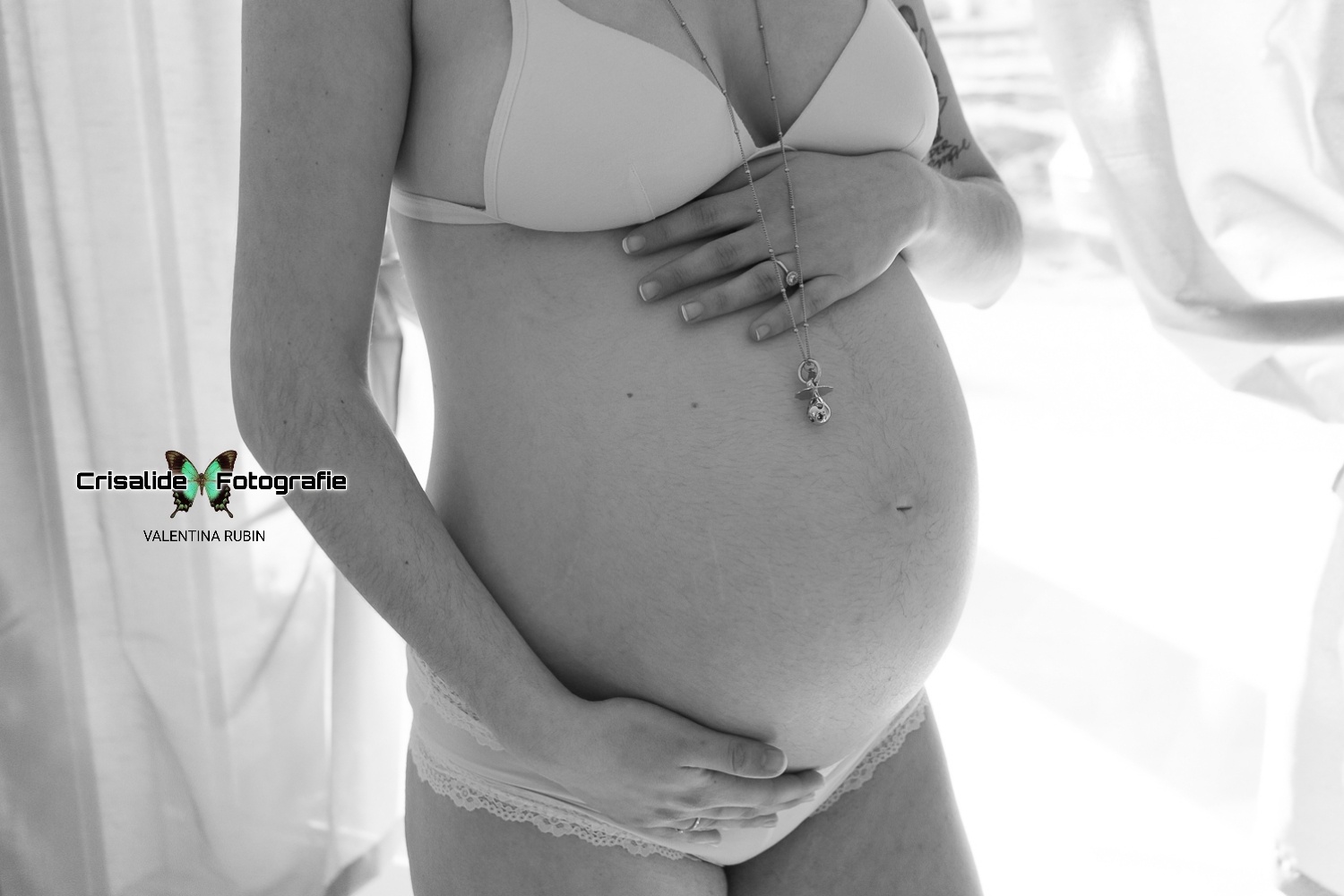 Scatto in bianco e nero. Primo piano del pancione girato di tre quarti, una mano sopra e una sotto, finestra con tenda sullo sfondo