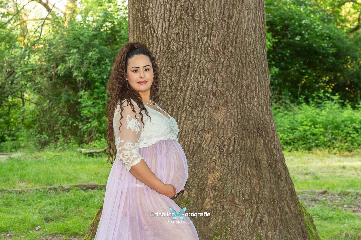Donna incinta di profilo con vestito bianco in pizzo e gonna in tulle violetto, che sorride all'obiettivo con le mani sotto la pancia, davanti a un tronco di quercia secolare e circondata da alberi e prato verde