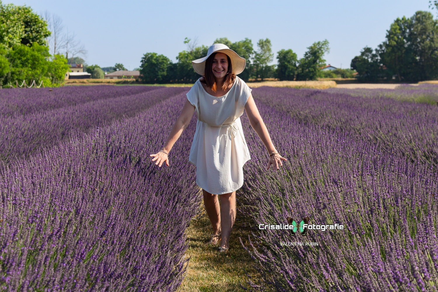 Mamma vestita di bianco con cappello, sorridente in un campo di lavanda sotto il cielo azzurro durante un servizio fotografico di famiglia