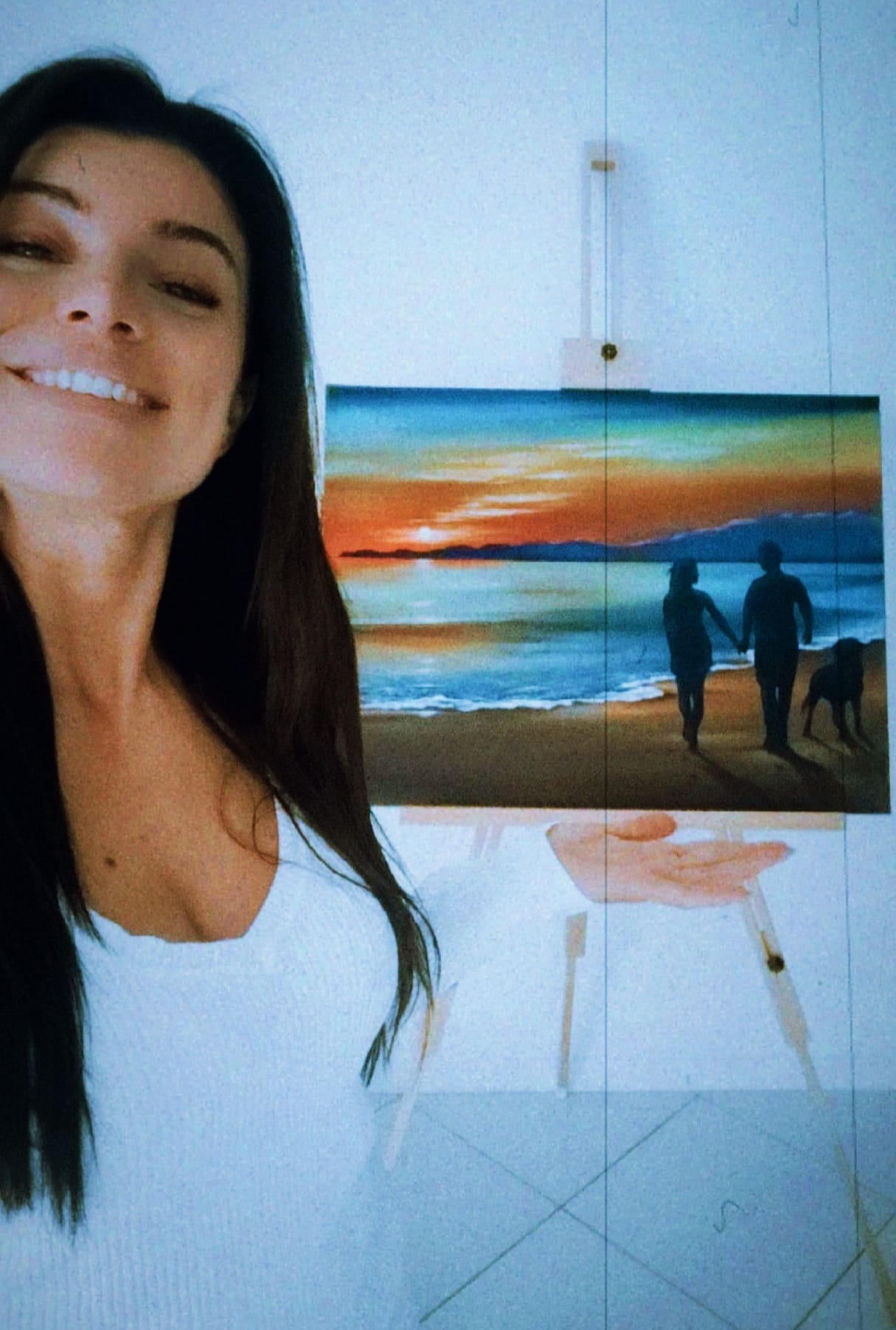 L'artista Jenny Corrò JeJe sorridente in primo piano mentre mostra un suo dipinto acrilico su tela che raffigura una coppia a piedi nudi sulla spiaggia al tramonto