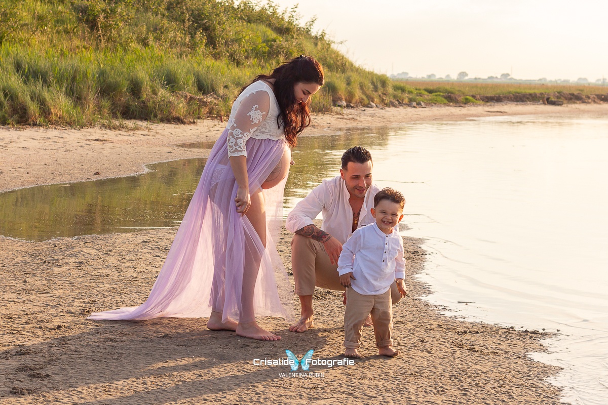 Famiglia in attesa sulla spiaggia: mamma incinta con vestito bianco e tulle violetto, papà accucciato che sorride al figlio di un anno e mezzo, e bambino sorridente che guarda l'obiettivo