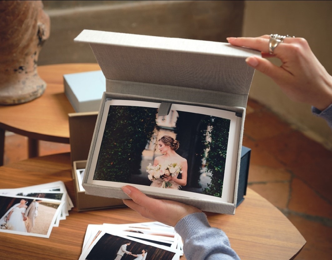 Cofanetto in cartone rivestito in tessuto, contenente stampe fotografiche 10x15 cm. Una mano di donna sorregge il cofanetto, l'altra apre il coperchio per mostrare le stampe all'interno. Sullo sfondo, tavolino con altre stampe appoggiate. Foto in luce naturale, dettaglio prodotto elegante e curato