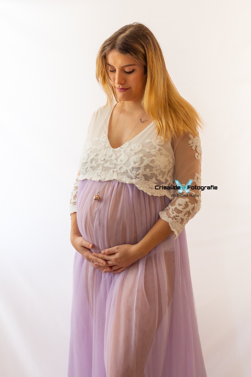 Mamma incinta con abito bianco in pizzo, manica tre quarti, gonna in tulle lilla, mani intrecciate sotto la pancia. Guarda il pancione. Ciondolo che scende delicatamente sul ventre. Capelli biondi sciolti sulle spalle. Sfondo bianco, atmosfera intima e dolce