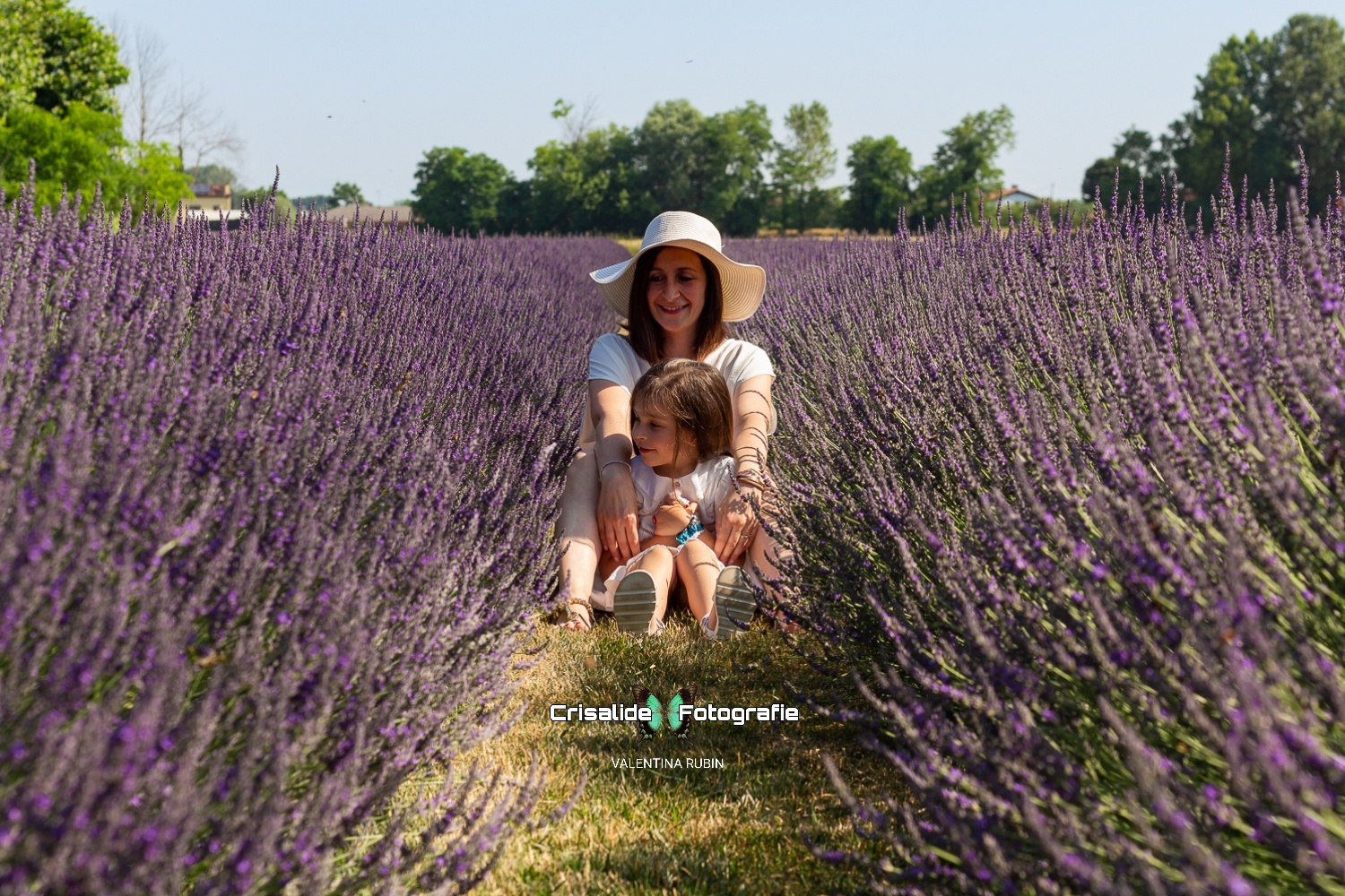 Mamma e figlia sedute sull'erba tra i filari di lavanda, entrambe vestite di bianco e sorridenti, mamma con le mani sulle spalle della bambina