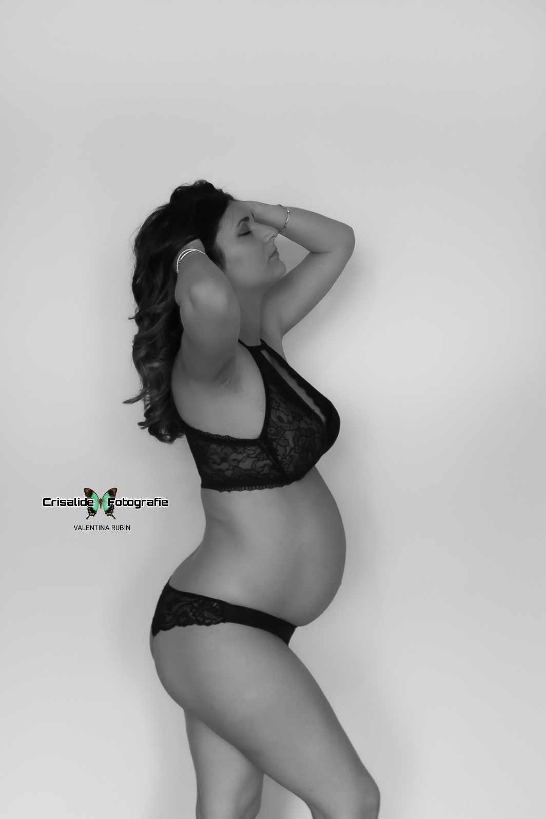 Mamma incinta in intimo nero, di profilo, testa rivolta verso l'alto, occhi chiusi e mani tra i capelli, gamba piegata