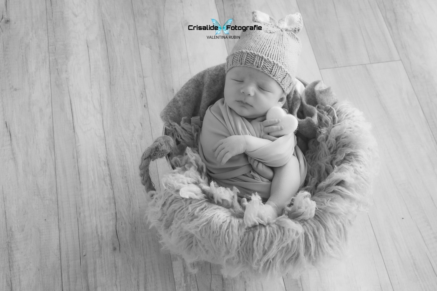 Neonato che dorme dentro un secchio, avvolto in fascia grigia con berretto da coniglietto, cuore bianco in mano, scatto newborn in bianco e nero