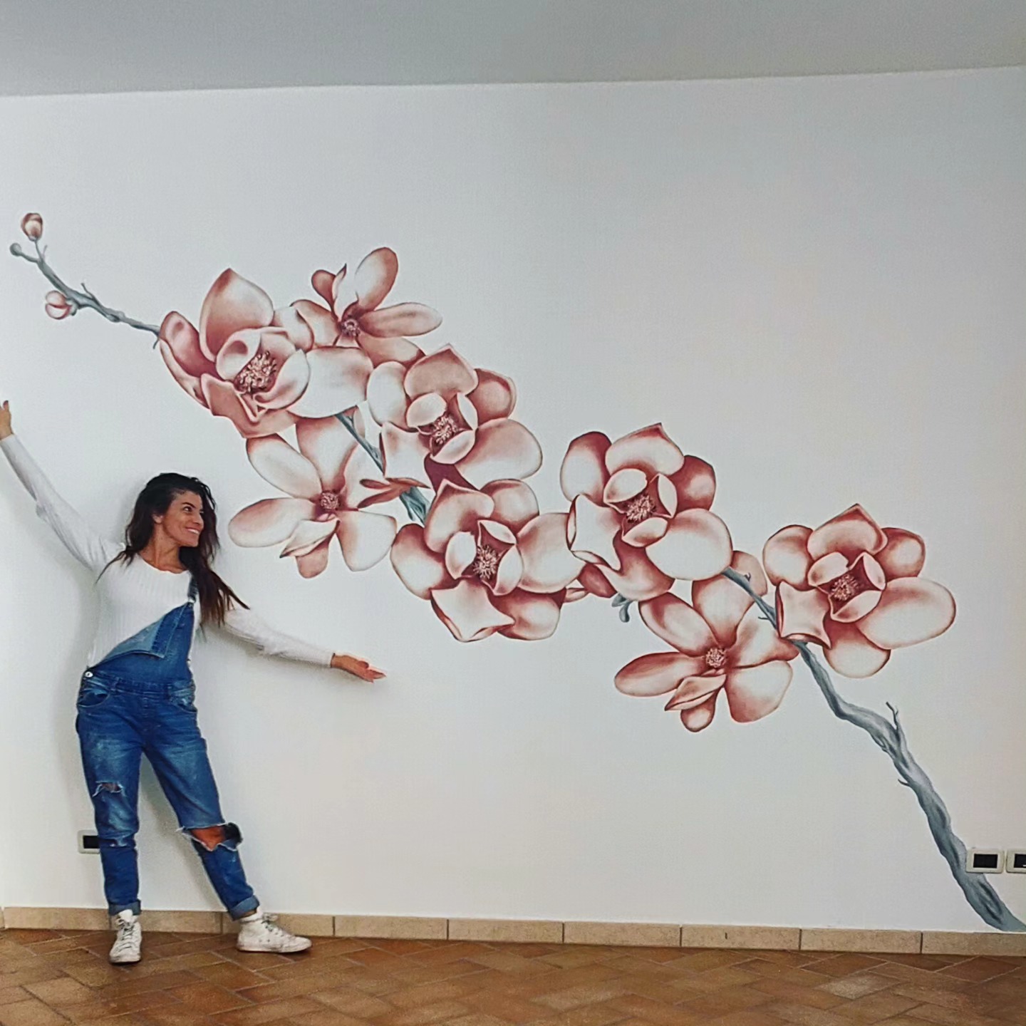L'artista Jenny Corrò JeJe sorridente a braccia aperte mentre mostra un grande dipinto murale da lei realizzato su una parete bianca, raffigurante un ramo con fiori rosa