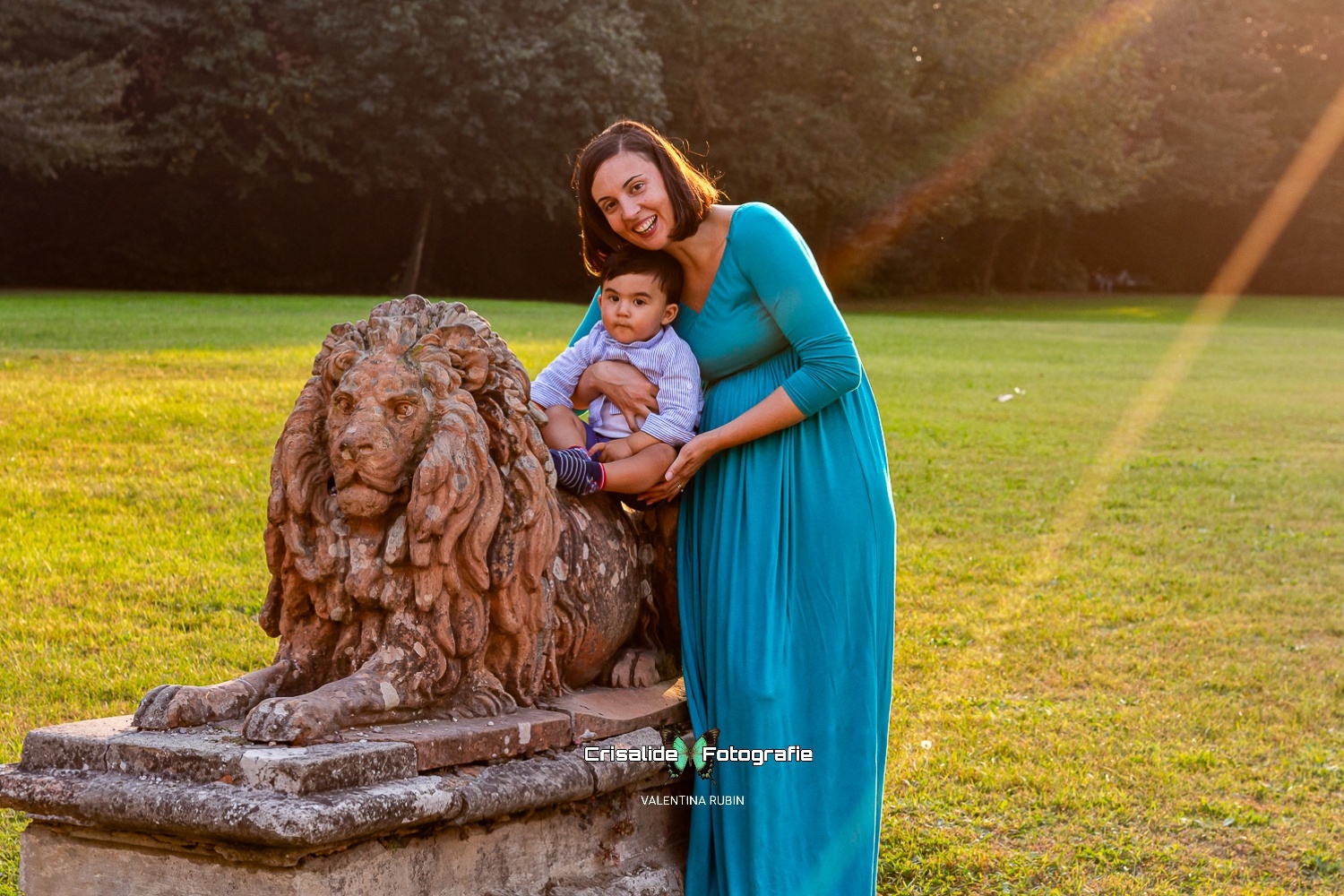 Mamma in piedi con abito lungo verde sorride all'obiettivo mentre tiene il suo bambino seduto su una statua di leone, con prato verde, alberi e un raggio di sole diagonale sullo sfondo