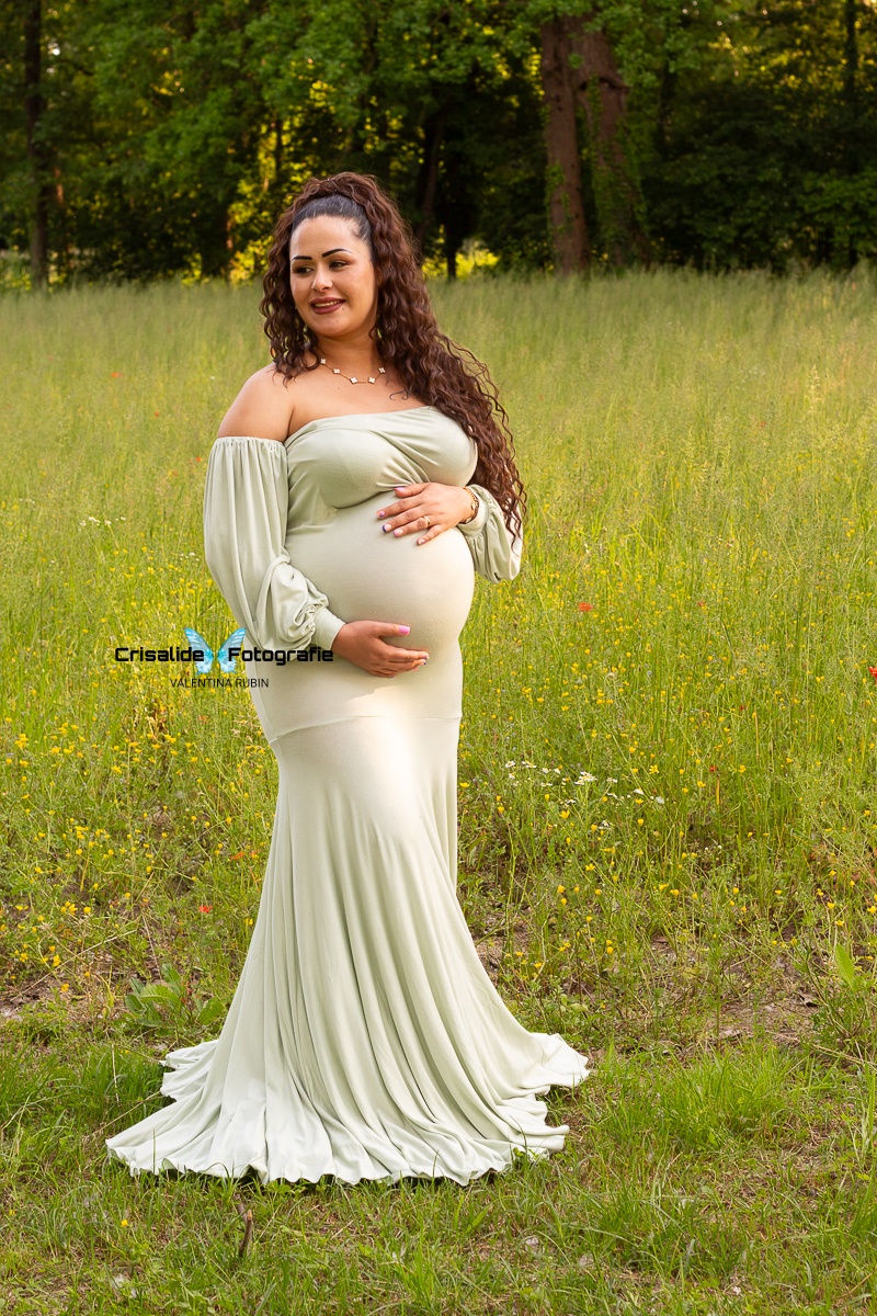 Mamma incinta in abito verde a sirena, tre quarti, sguardo laterale e sorriso, mani sul pancione, capelli lunghi mori ondulati, sfondo di prato con fiori gialli e alberi verde scuro