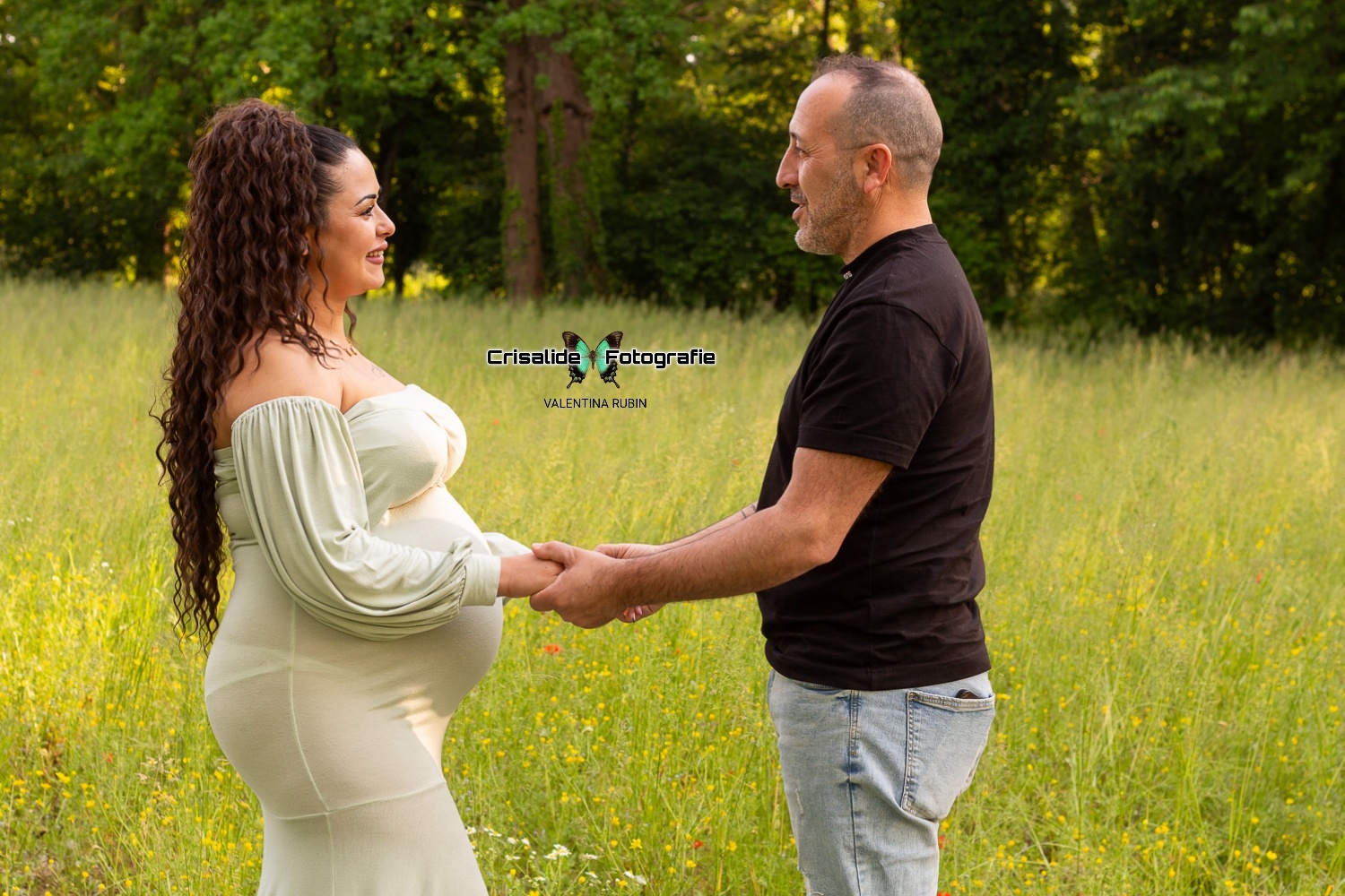 Donna incinta con vestito verde chiaro a sirena che sorride al marito, entrambi di profilo, mano nella mano su un prato con alberi sullo sfondo