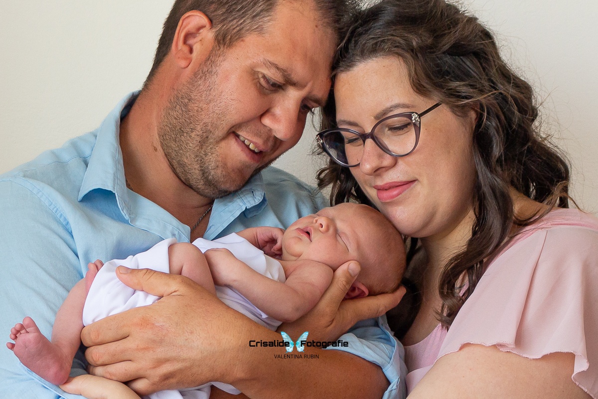 Papà in camicia azzurra tiene la neonata fasciata di bianco mentre la mamma in abito rosa appoggia la testa a quella del marito, i tre volti formano un triangolo su sfondo bianco