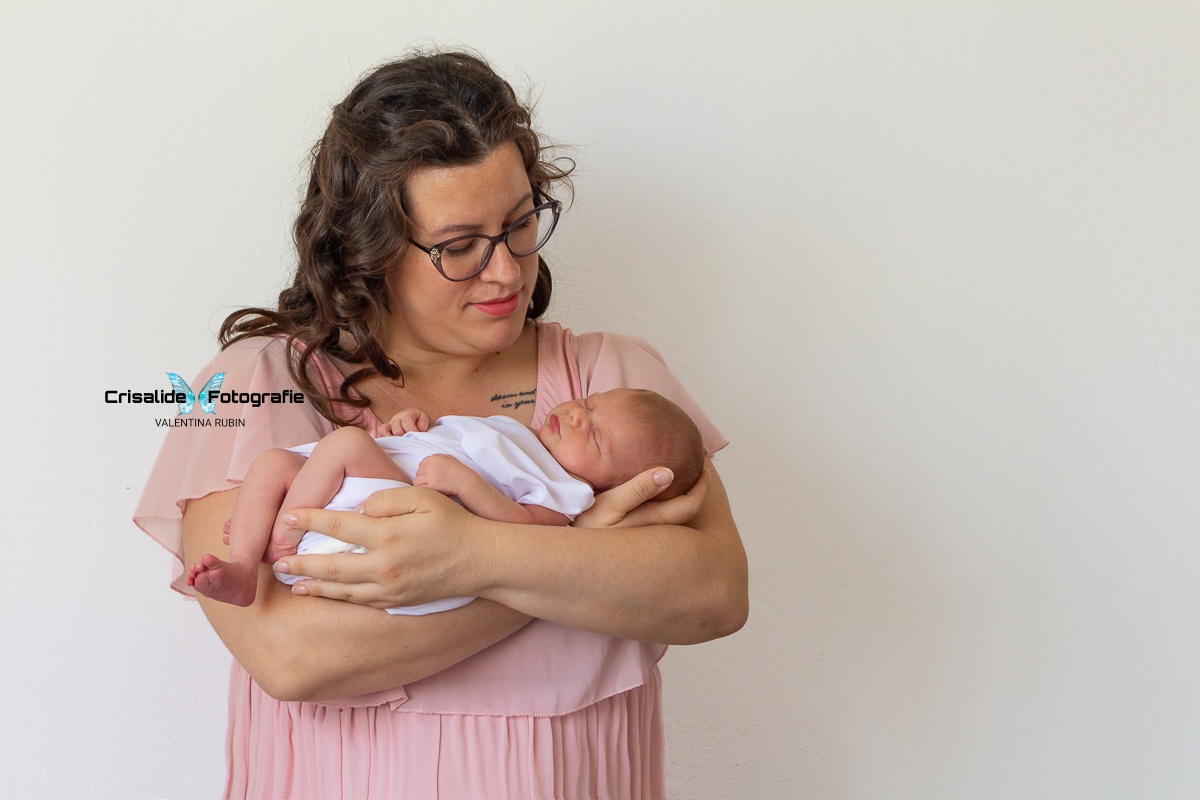 Mamma con vestito in chiffon rosa a maniche corte, tiene in braccio una neonata fasciata di bianco. La mamma, con occhiali e capelli castani a boccoli sulle spalle, guarda la bambina sorridendo. La neonata dorme. Sfondo completamente bianco