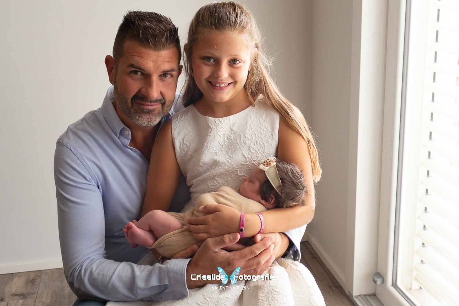 Papà e bambina sorridenti guardano l'obiettivo mentre tengono in braccio la neonata che dorme, luce naturale da finestra, sfondo bianco