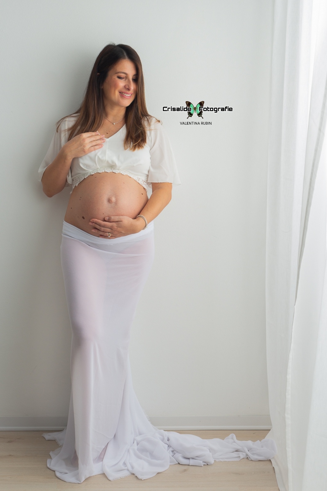 Mamma incinta in abito bianco con gonna in chiffon, ritratta in luce naturale accanto a una tenda mentre sorride guardando verso la finestra