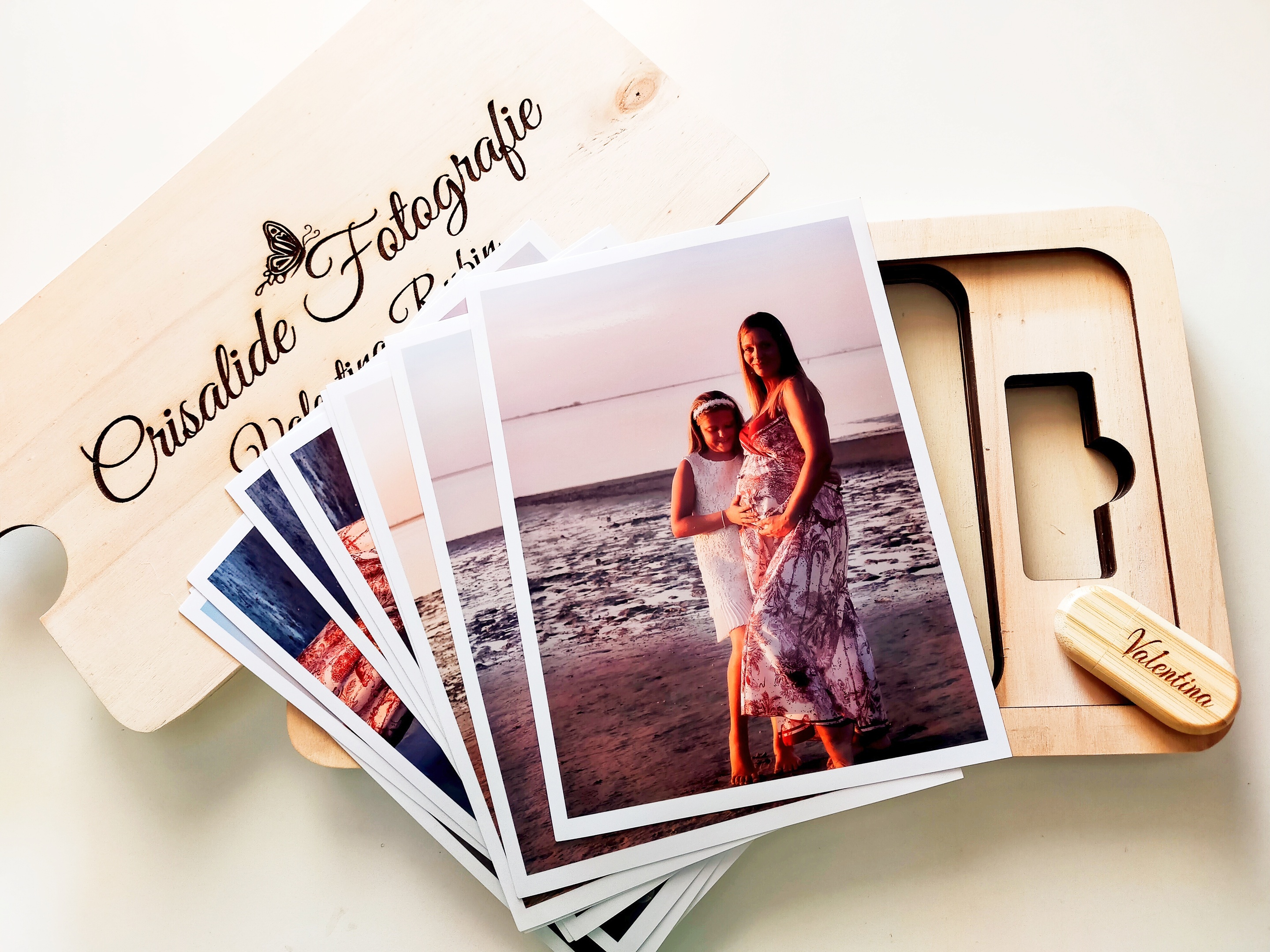 Memory box in legno con coperchio, posizionato a sinistra con logo inciso a laser. Al centro è disposto un ventaglio di stampe fotografiche; la prima mostra una mamma incinta insieme alla figlia di 7 anni su una spiaggia. Sulla destra è presente una chiavetta USB in legno con nome inciso a laser. Sfondo completamente bianco