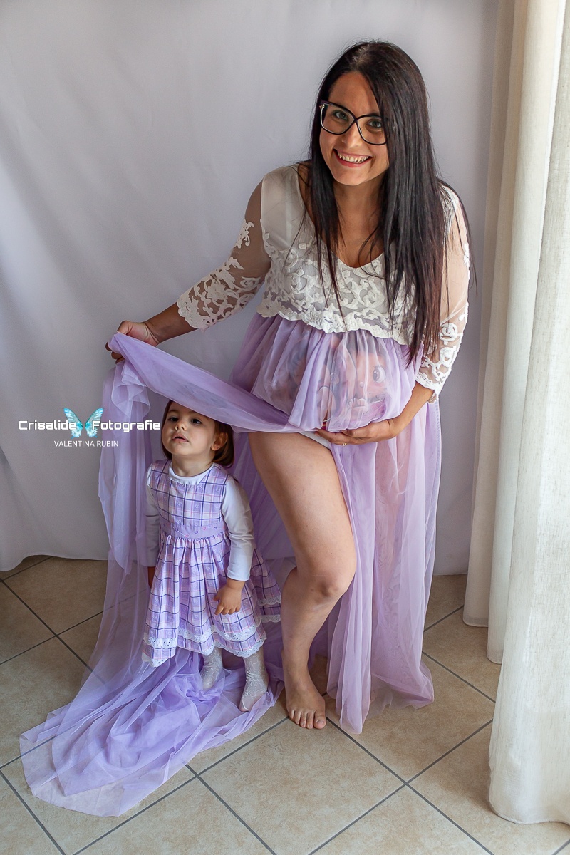 Mamma incinta in abito bianco di pizzo con gonna lilla, di fronte all'obiettivo mentre sorride, accanto la figlia di 2 anni in vestito lilla che guarda verso l'alto coperta dalla gonna, sfondo bianco con tenda laterale