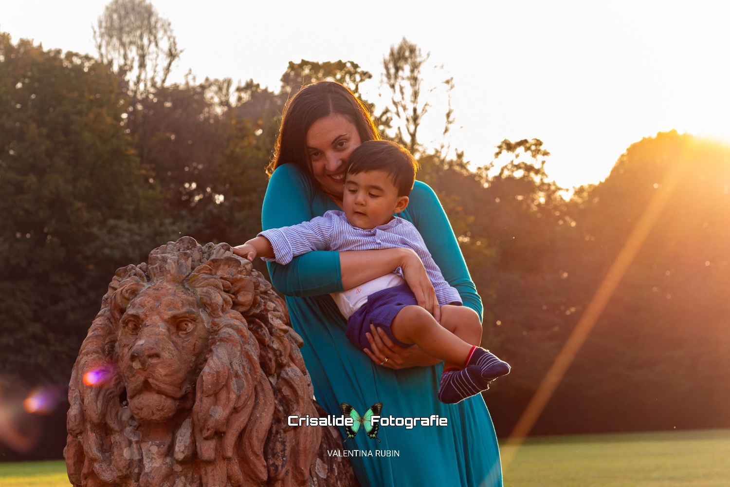 Mamma sorridente in abito lungo verde tiene in braccio il figlio con pantaloncini blu e camicia a righe azzurre; il bambino tocca la testa di una statua di leone, un raggio di sole illumina la scena