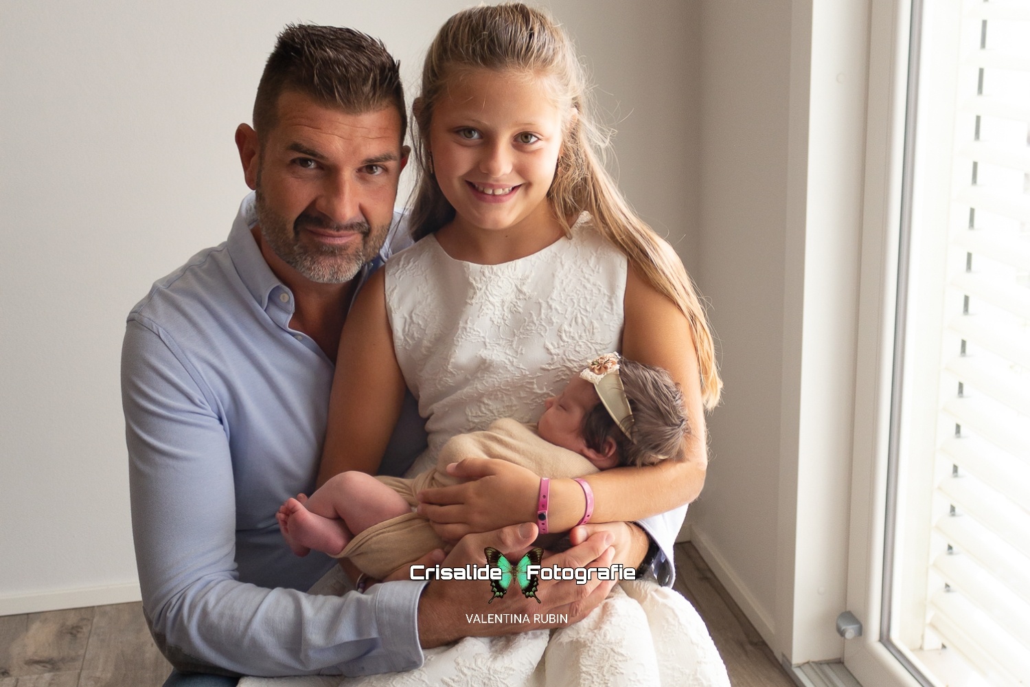 Papà e bambina sorridenti guardano l'obiettivo mentre tengono in braccio la neonata che dorme, luce naturale da finestra, sfondo bianco