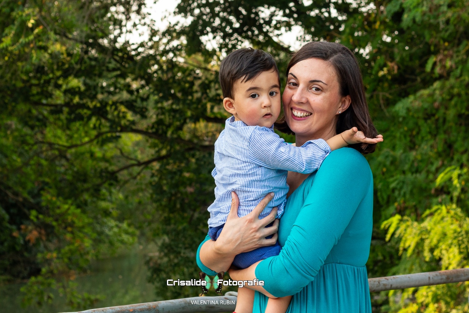Mamma con abito verde tiene in braccio il suo bambino di 10 mesi, entrambi guardano l'obiettivo sorridenti, con alberi e fiume sullo sfondo