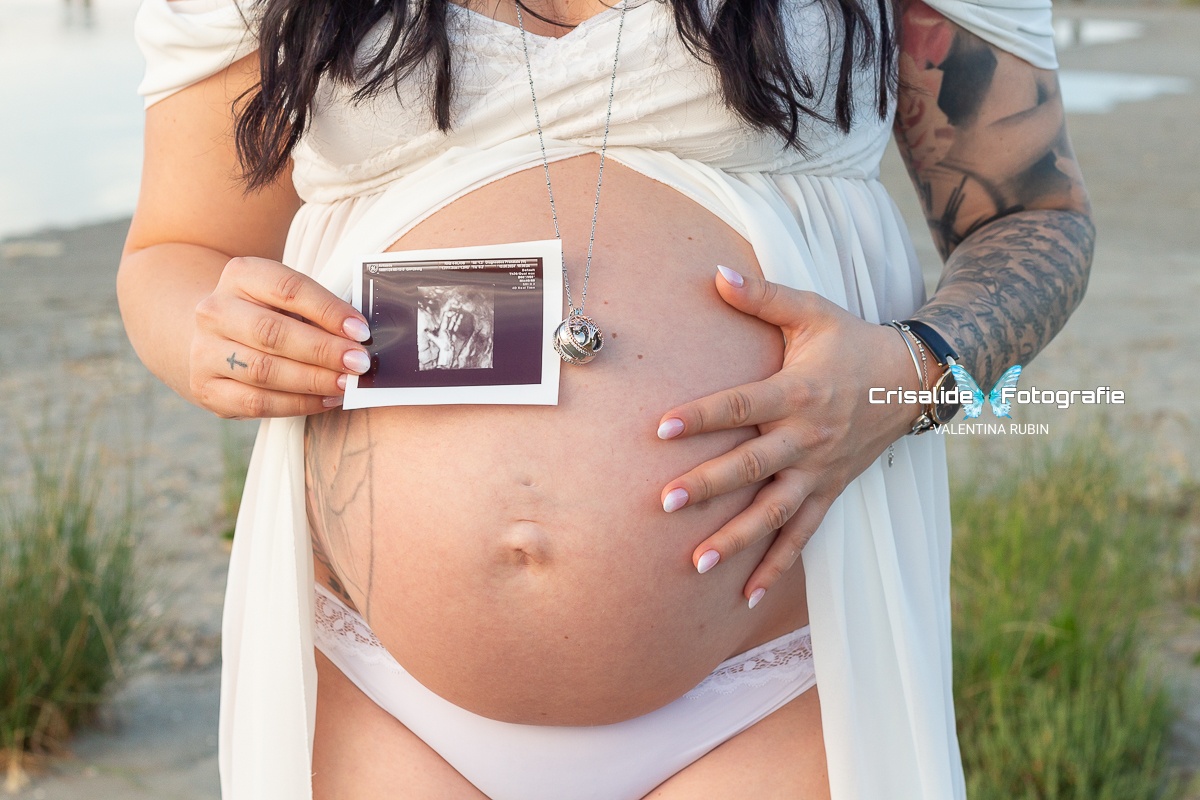 Primo piano del pancione di mamma in abito bianco di chiffon, mano sul pancione e nell'altra mano l'ecografia, ciondolo sulla pancia, sfondo spiaggia e mare