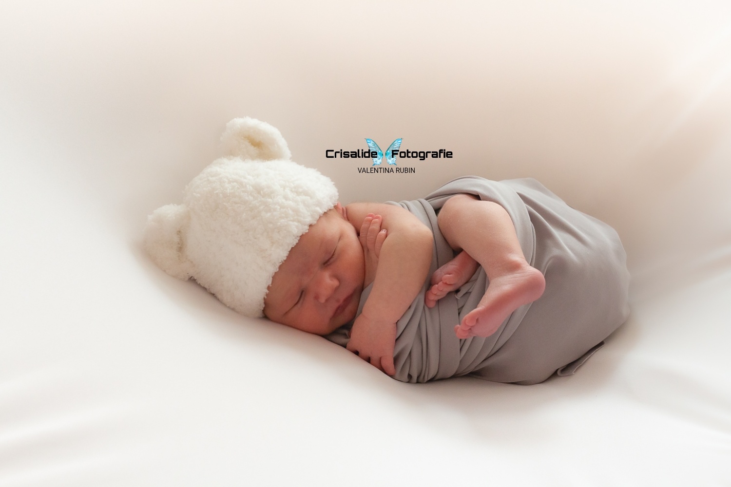 Neonato che dorme girato di lato, fasciato in grigio con berretto bianco e orecchiette da orsacchiotto, sfondo bianco