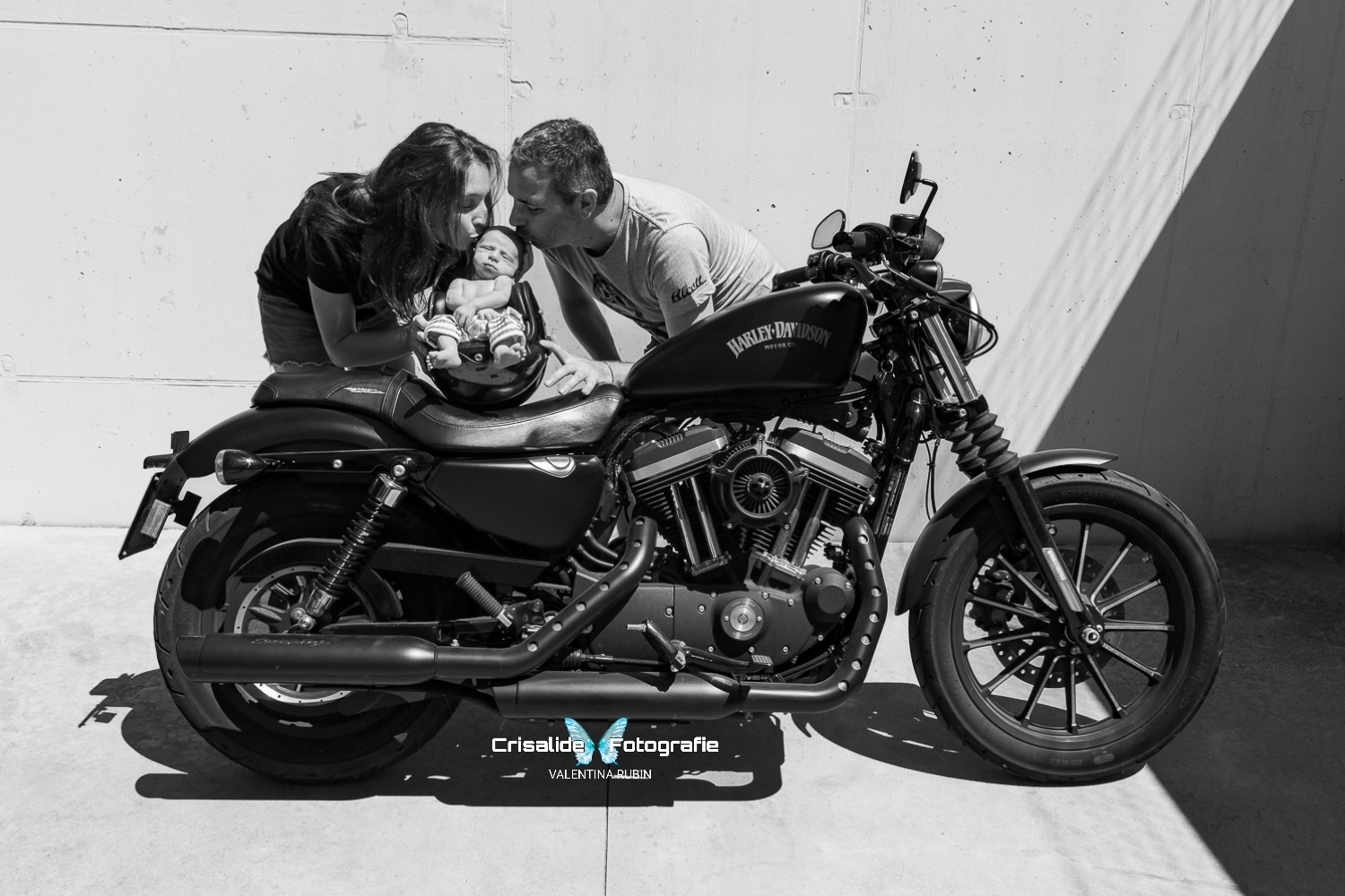 Mamma e papà baciano la testina del neonato che dorme dentro un casco appoggiato su una moto Harley Davidson, scatto newborn in bianco e nero