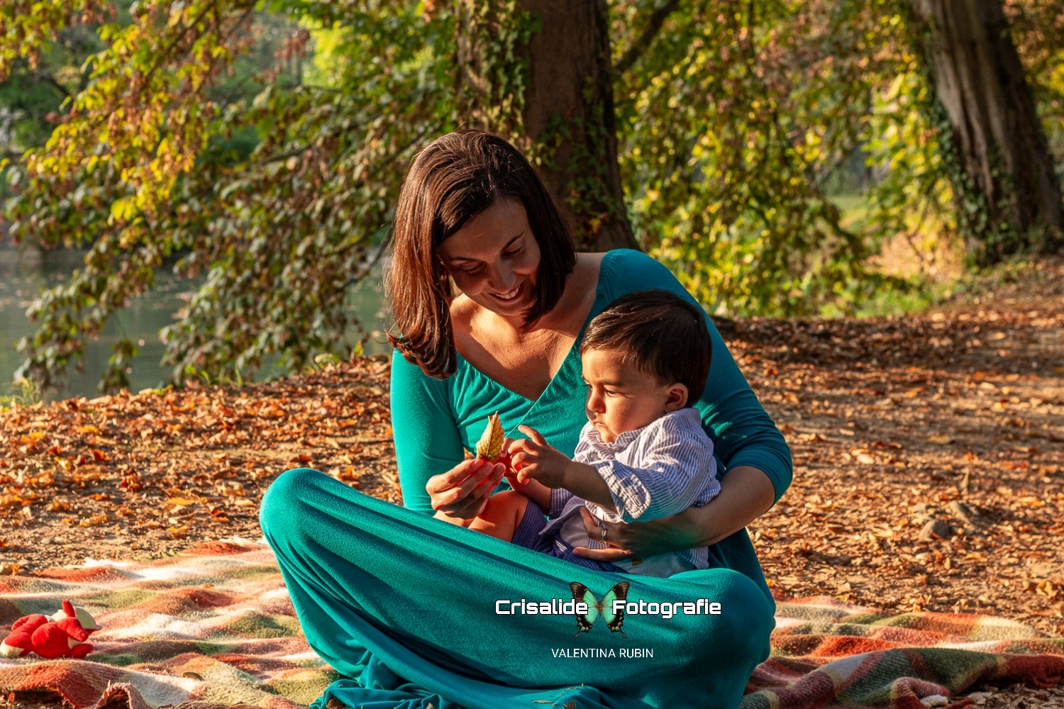 Mamma seduta su una coperta tiene in braccio il suo bambino mentre gli mostra una foglia secca, con alberi e foglie autunnali sullo sfondo