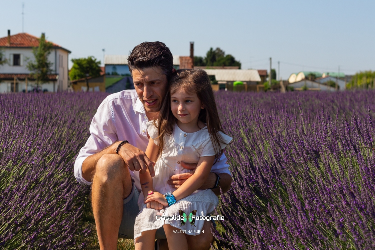 Papà accucciato con la figlia in braccio, entrambi vestiti di bianco, sorridono guardando di lato tra i filari di lavanda