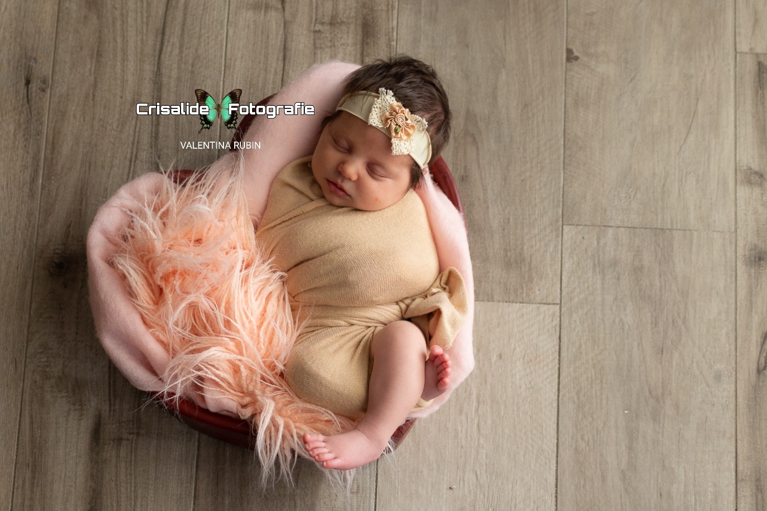 Neonata che dorme dentro ad una cestina a forma di cuore, con lana e pelliccia rosa, fasciata di tessuto beige, coroncina con fiocchetto in testa, sfondo pavimento beige