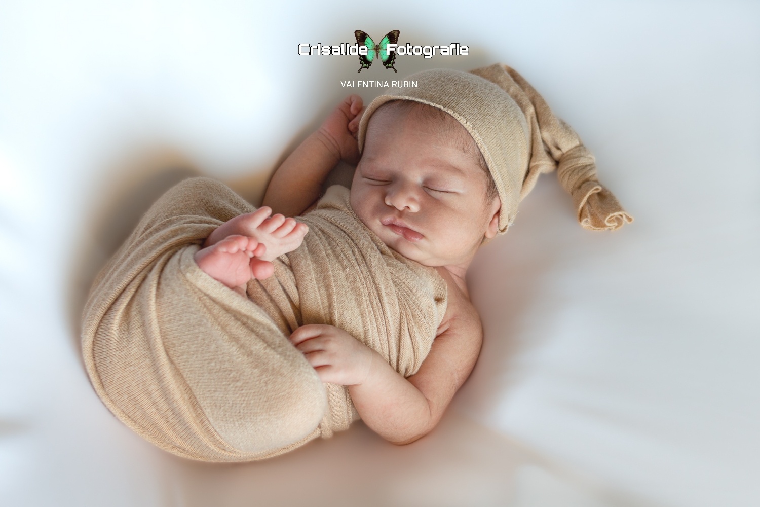 Neonato che dorme, fasciato di beige, con berrettino coordinato, ha una manina vicino la testa, sfondo bianco