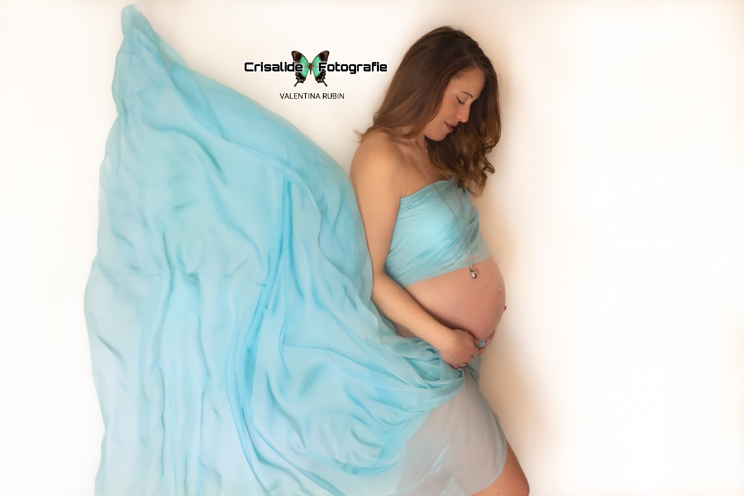 Mamma incinta di profilo avvolta in un telo di chiffon azzurro svolazzante su sfondo bianco, ritratto delicato di maternità