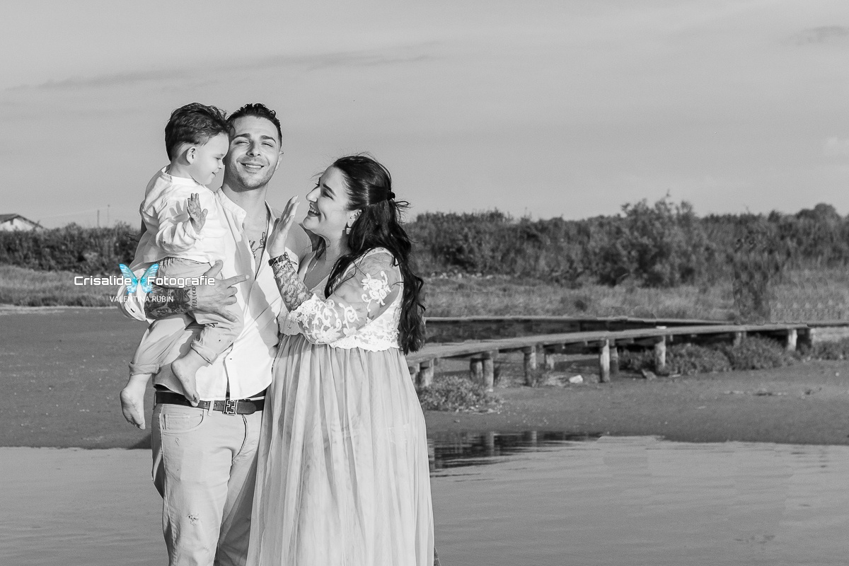 Mamma in abito bianco di pizzo e papà in camicia bianca e pantaloni beige, tiene in braccio il bambino sorridente pronto a dare il cinque alla mamma, sfondo pontile, mare e vegetazione
