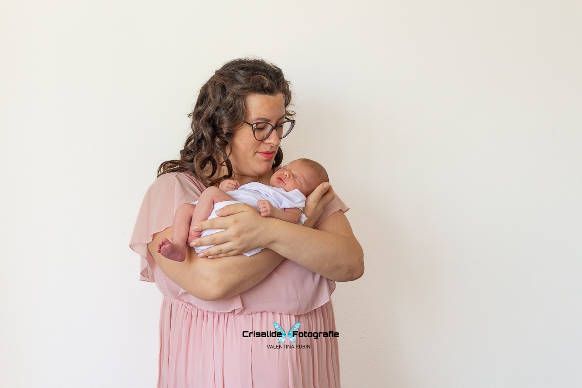 Mamma in abito di chiffon rosa che guarda sorridendo la neonata addormentata tra le sue braccia su sfondo bianco