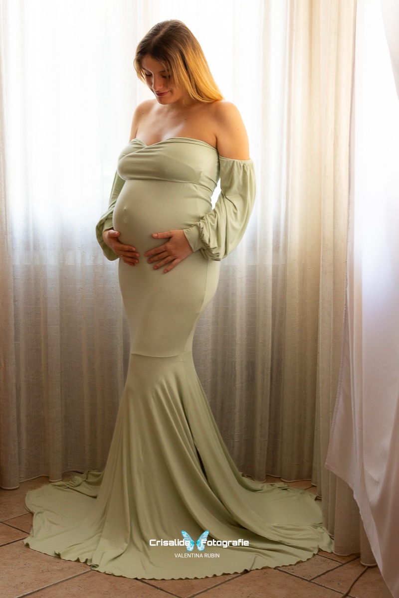 Ritratto di una donna incinta che accarezza la pancia con un vestito verde chiaro, illuminata dalla luce delle finestre nella sua casa