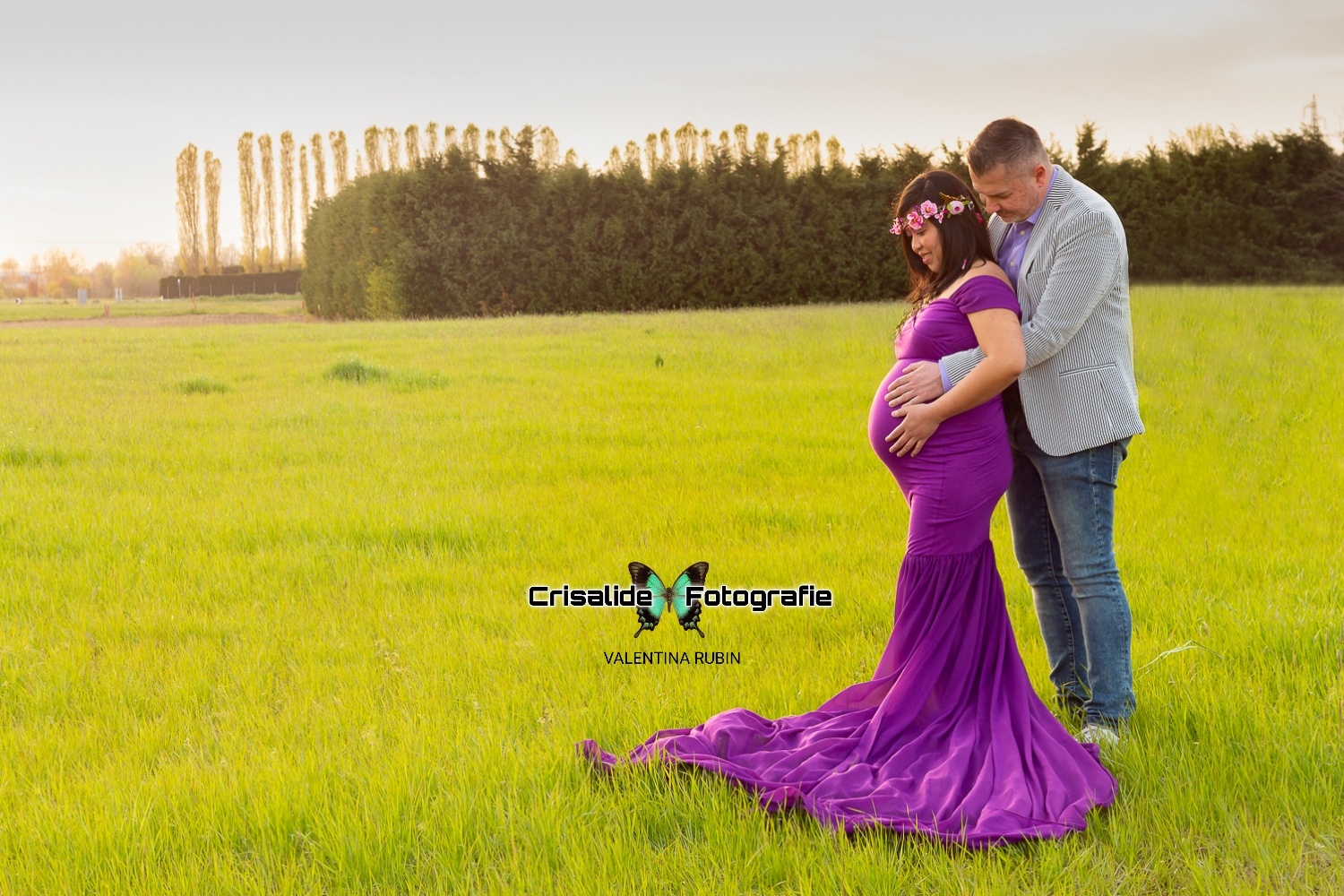 Mamma incinta con papà alle spalle che la abbraccia, entrambe le mani sul pancione, guardando la pancia, sul prato verde chiaro con alberi sullo sfondo
