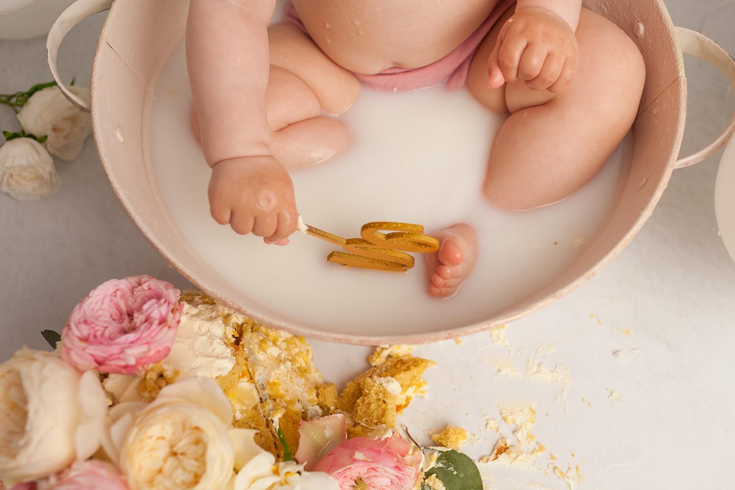 Bambina di 1 anno, con body bianco e gonna in tulle rosa, seduta a terra con davanti un cupcake con la candelina accesa. Di fianco un numero 1 e cubi con scritta one. Sfondo con bandierine colorate e palloncini