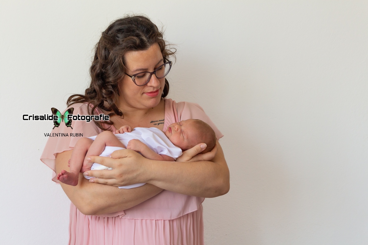 Mamma con vestito in chiffon rosa a maniche corte, tiene in braccio una neonata fasciata di bianco. La mamma, con occhiali e capelli castani a boccoli sulle spalle, guarda la bambina sorridendo. La neonata dorme. Sfondo completamente bianco