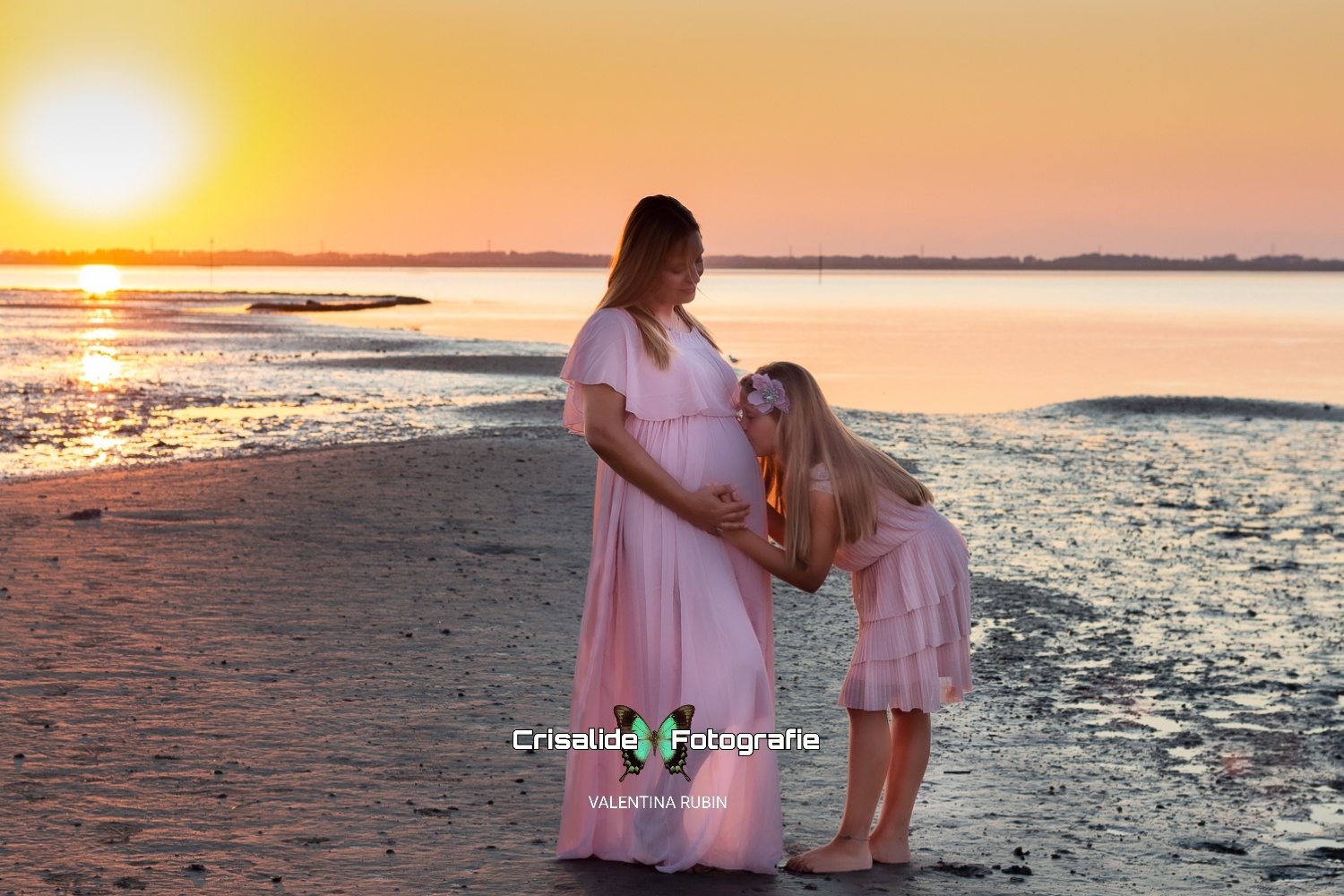 Mamma incinta in abito rosa sulla spiaggia con la figlia di 8 anni, anche lei vestita di rosa. La bambina bacia la pancia della mamma e appoggia una mano sopra, mentre la mamma sorride e poggia la sua mano sopra quella della figlia. Capelli lisci della mamma sulle spalle, capelli biondi della bambina con fiore tra i capelli. Sfondo mare con riflessi dorati del sole all'orizzonte