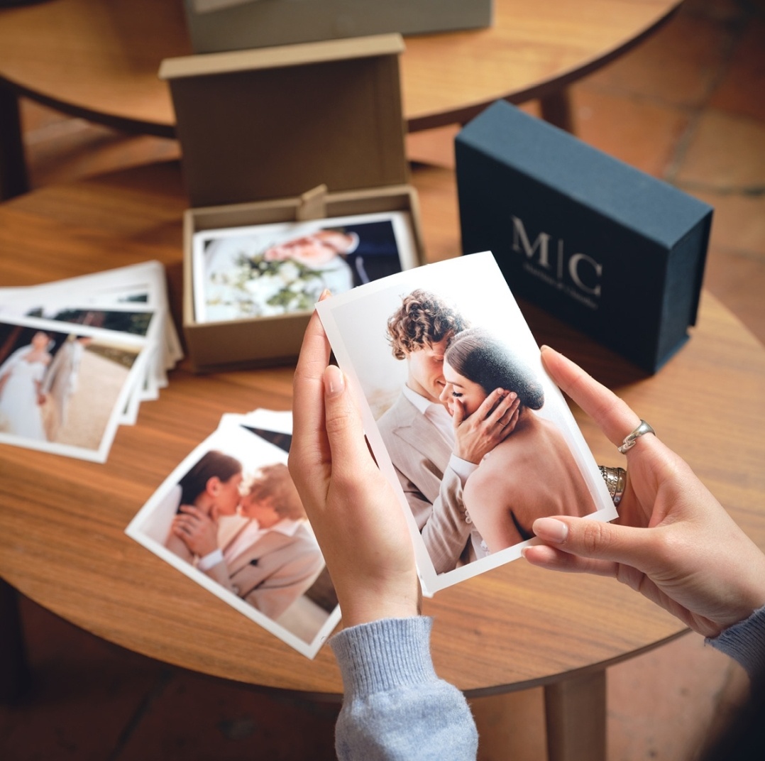 Stampa fotografica newborn raffigurante una coppia, due mani che sorreggono la stampa, sfondo tavolino con altre stampe appoggiate, box in cartone con coperchio rivestito in tessuto blu personalizzato con logo, contenente 20 stampe 10x15 cm - Pacchetto Maxi con servizio newborn a domicilio