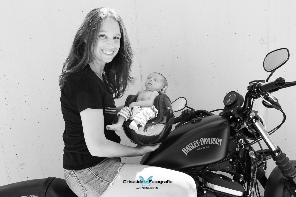 Mamma sorridente seduta sulla sua moto Harley Davidson che guarda in camera mentre tiene il casco con il suo bambino seduto all'interno, in un ritratto in bianco e nero su sfondo bianco