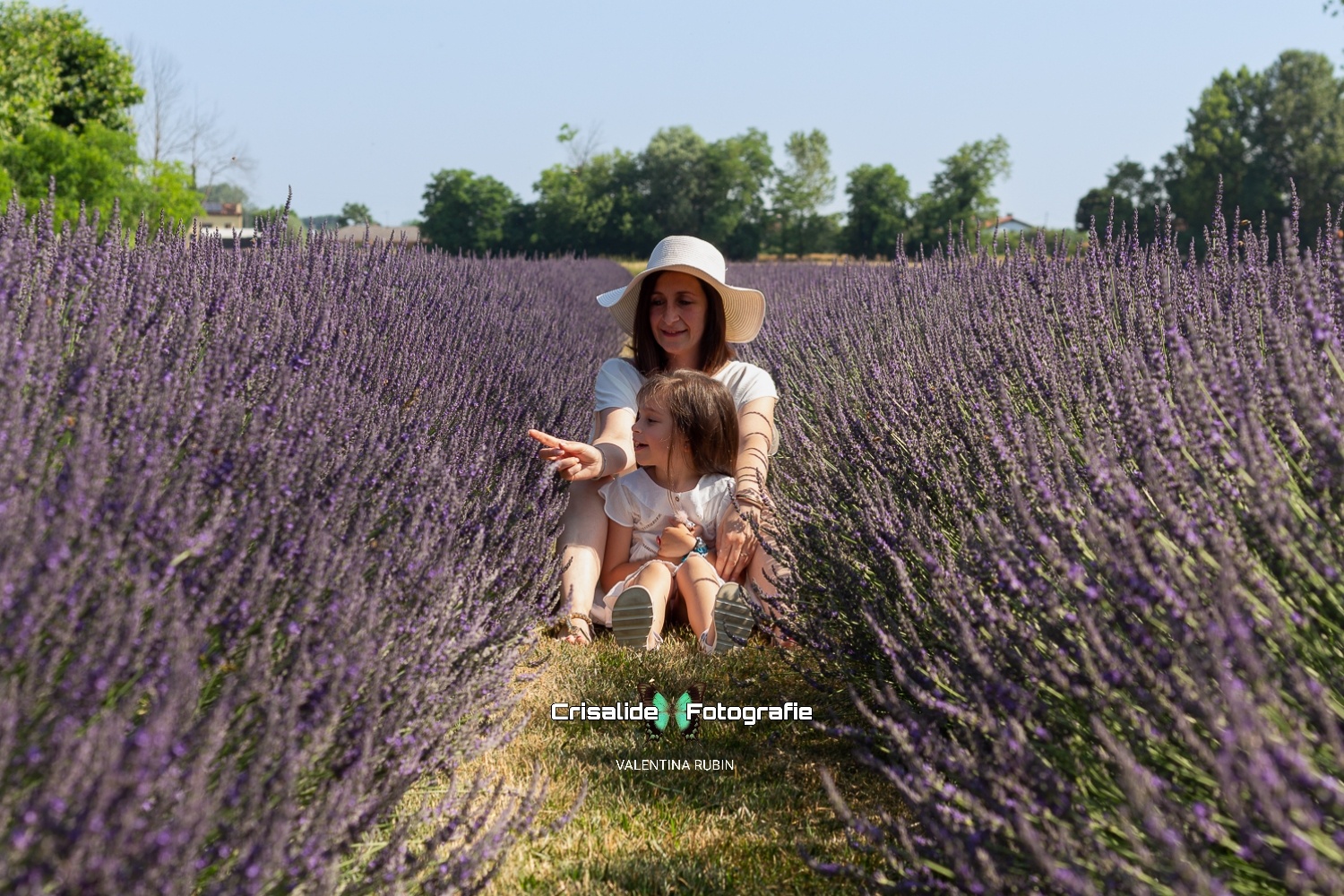 Mamma e figlia sedute tra i filari di lavanda, entrambe vestite di bianco, mamma indica la lavanda con il dito mentre rassicura la bambina preoccupata per le api