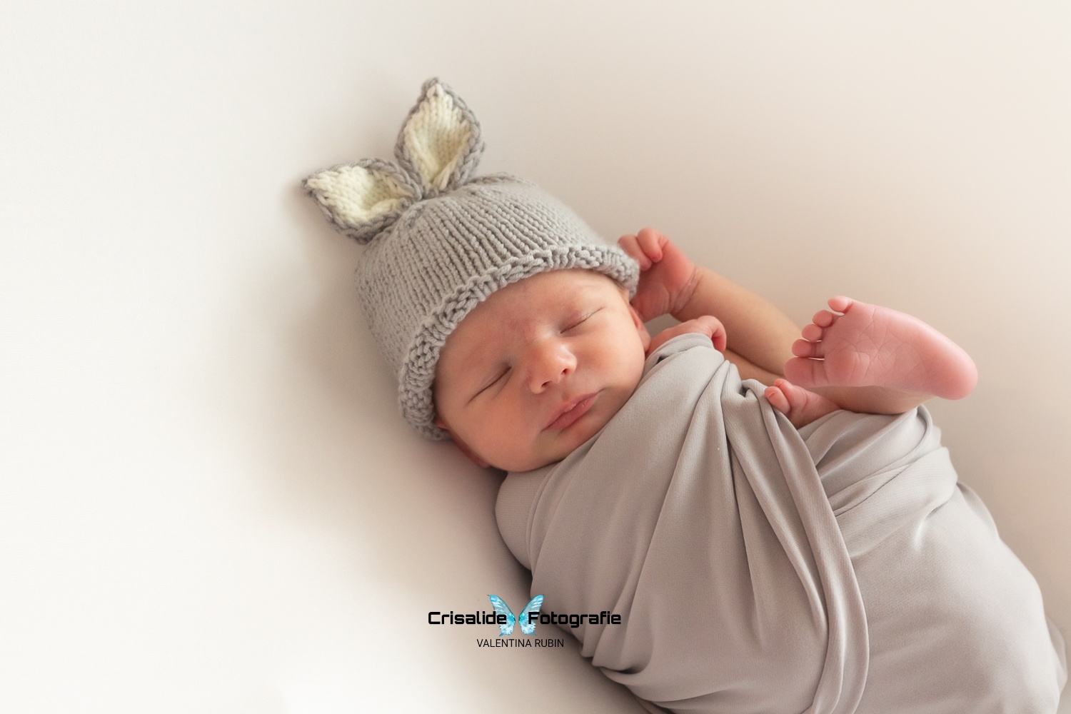 Neonato che dorme fasciato in fascia grigia, con piede alzato e manina dietro l'orecchio, berretto con orecchiette di coniglio, sfondo bianco