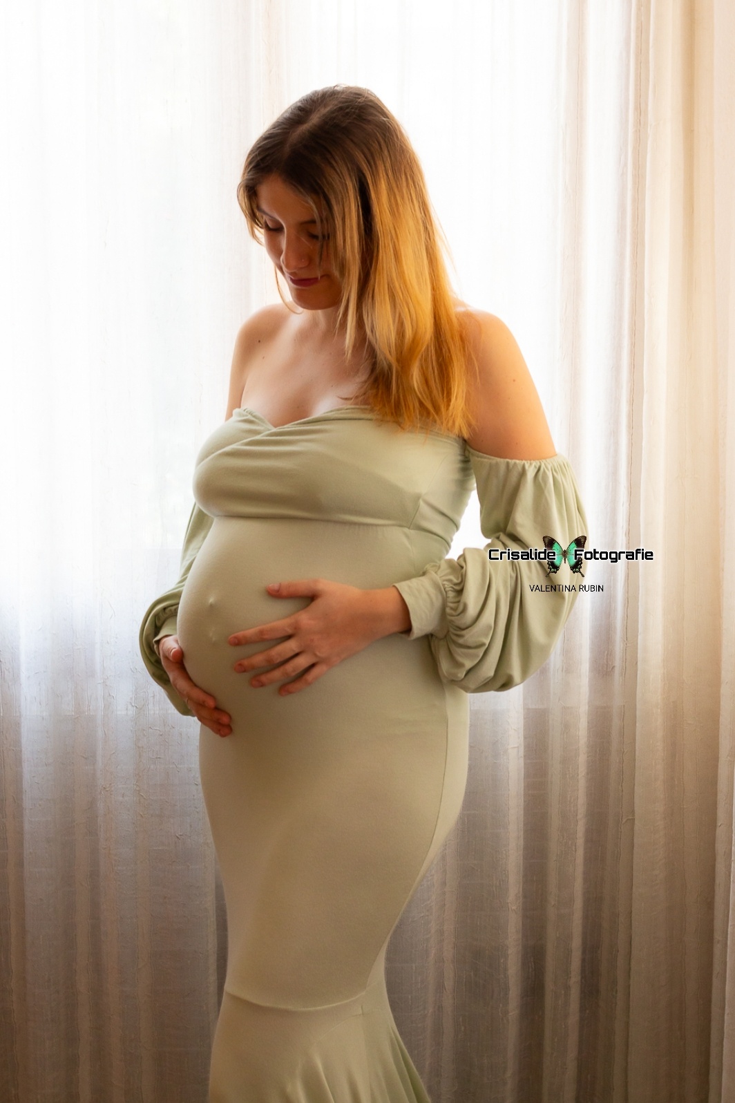 Mamma incinta in abito verde a sirena, girata di tre quarti mentre guarda il pancione sorridendo, luce dalla finestra dietro la tenda