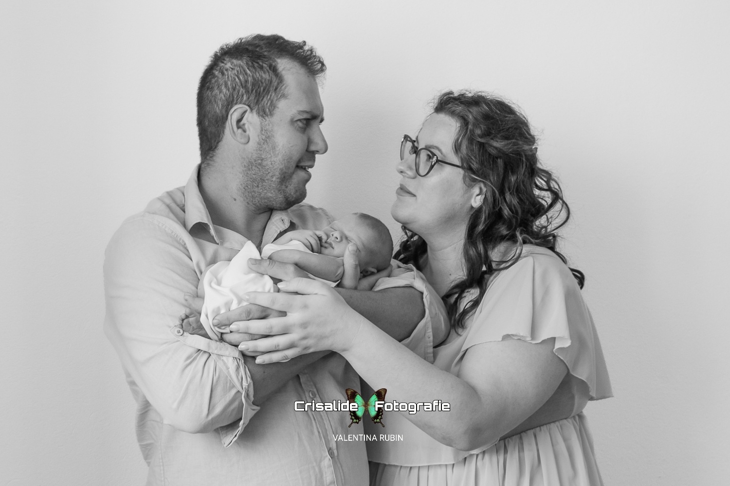Mamma e papà si guardano mentre tengono in braccio la neonata che dorme, fasciata in bianco, fotografia newborn in bianco e nero