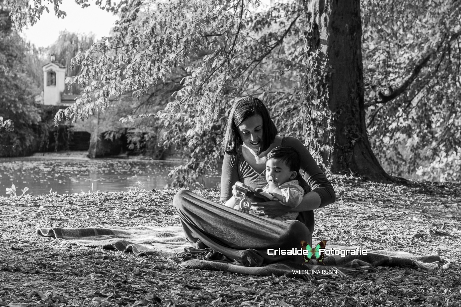 Mamma seduta su una coperta che tiene in braccio il suo bambino e lo distrae con una foglia secca, in un paesaggio autunnale in bianco e nero