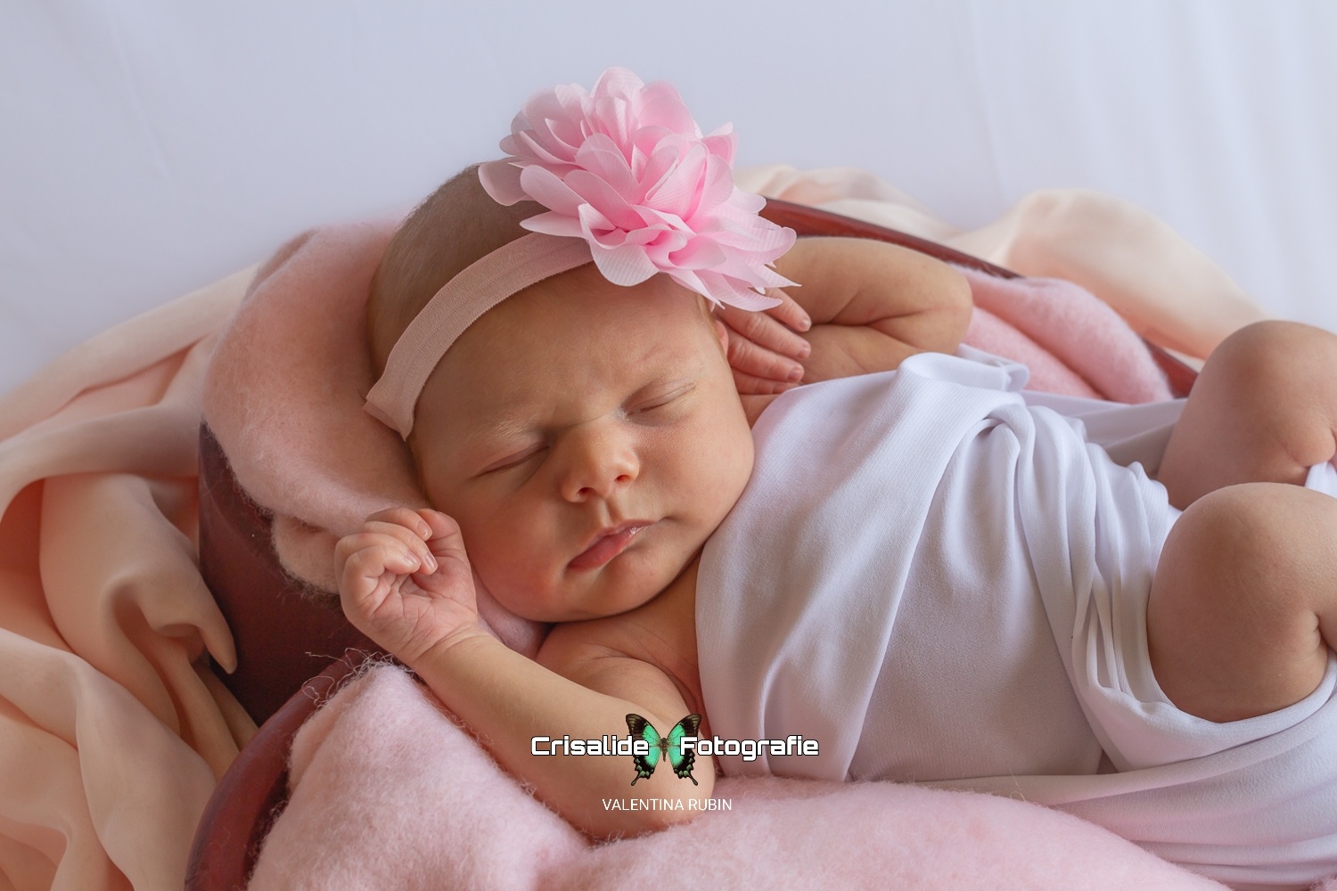 Neonata che dorme in una cestina a forma di cuore, fasciata di bianco, con fiore rosa sulla testa e tessuti rosa su sfondo bianco