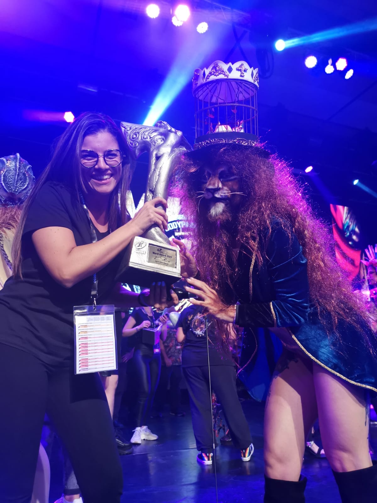 L'artista Jenny Corrò JeJe sorridente sul palco mentre solleva il trofeo del secondo posto al World Bodypainting Festival 2023, accanto al suo modello con un trucco totale da leone e una gabbia come copricapo