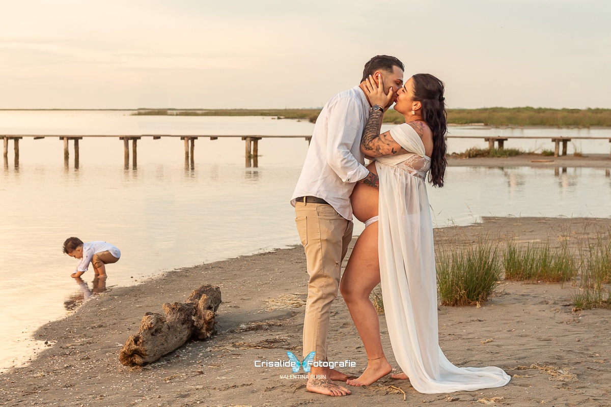 Mamma in chiffon bianco e marito in camicia bianca si baciano, bambino piccolo gioca accovacciato sulla sabbia, sfondo spiaggia, mare, pontile e ciuffi d'erba