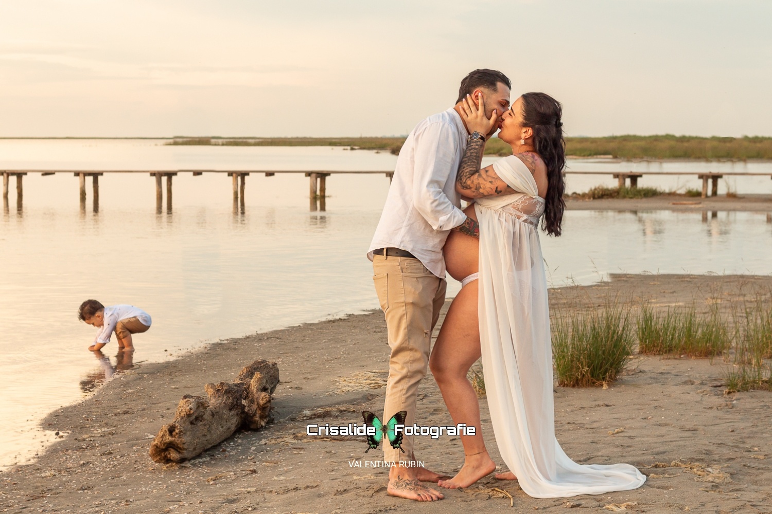 Mamma in chiffon bianco e marito in camicia bianca si baciano, bambino piccolo gioca accovacciato sulla sabbia, sfondo spiaggia, mare, pontile e ciuffi d'erba